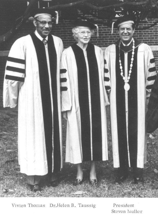 Vivien Thomas (left) together with Dr. Helen B. Taussig (middle) and President Steven Muller (right) are smiling in an old photograph with a brick wall in their background. Vivien is wearing an eyeglass and academic gown, a hat, and black shoes so as Helen, while Steven is wearing a long medal and an academic gown, hat and black shoes