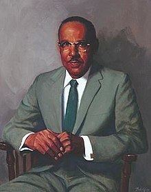 Vivien Thomas sitting on a wooden chair while holding his hands together with a mustache, wearing an eyeglass and a white long sleeve with a dark green necktie under his light green coat