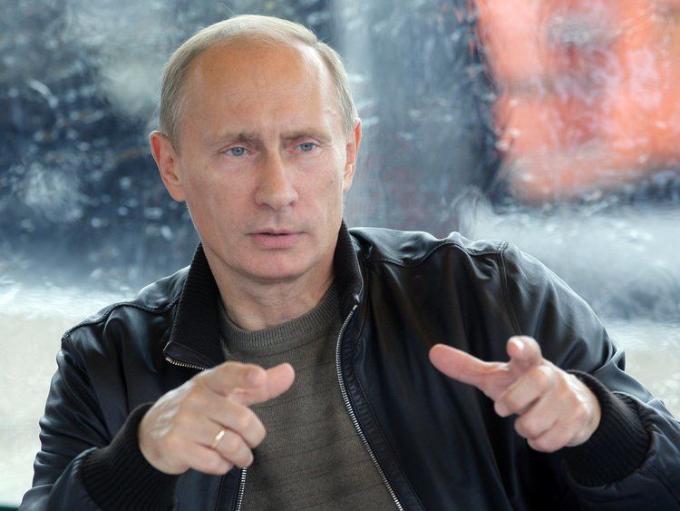 Vladimir Putin with a serious face while moving his hands, wearing a black jacket over a gray shirt and a ring.
