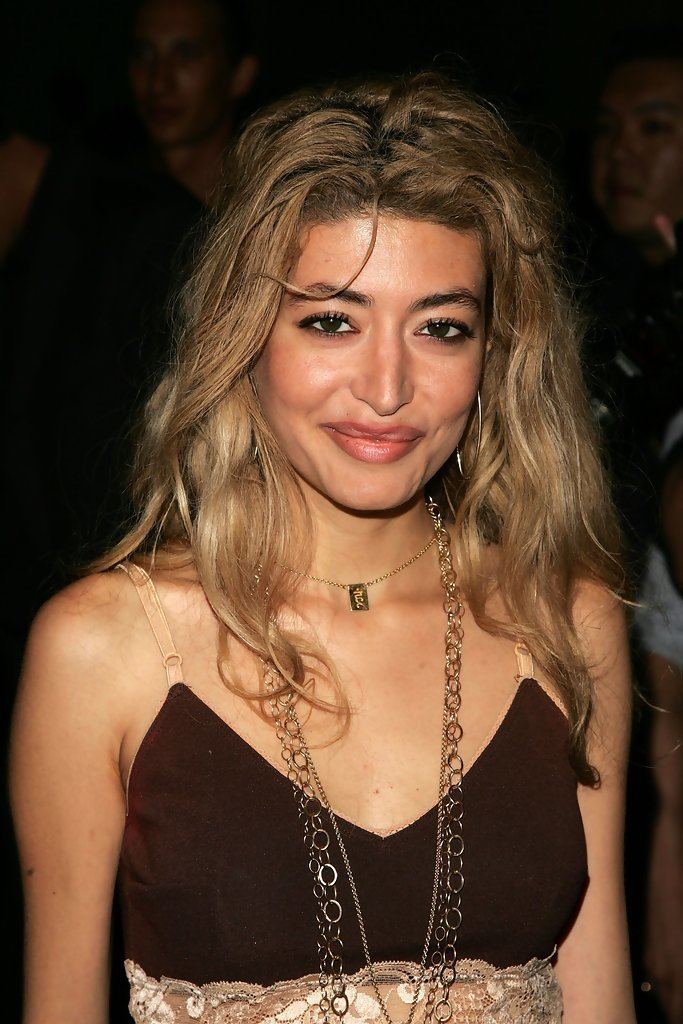 Wafah Dufour smiling, with curly blonde hair, wearing hoop earrings, necklaces, and a brown spaghetti top.