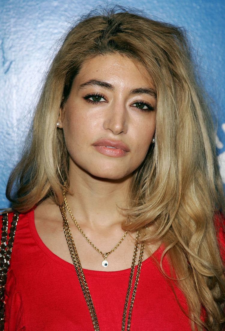 Wafah Dufour with a serious face, with wavy blonde hair, wearing pearl earrings, necklaces, and a red shirt.