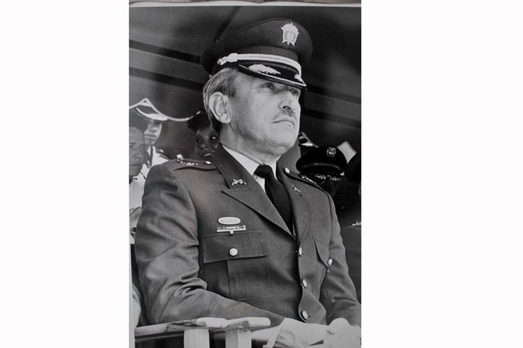 Waldemar Franklin Quintero sitting on the chair while looking afar and wearing a police coat, long sleeve, necktie, and peaked cap