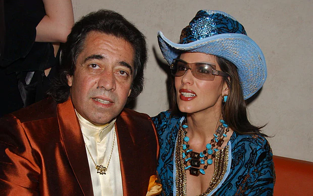In a room gray wall, with a woman standing, wearing a sleeveless black dress, in front, from left, Walid Juffali is serious, sitting on orange couch, has black hair wearing a gold necklace a cream long sleeve polo under a shiny brown coat with orange handkerchief on his left chest. At the right Christina Estrada is smiling, sitting leaning to her right, has brown long hair wearing a blue green fancy cowboy hat with, large tree layer beads necklace, cyan earrings, a gray-translucent sunglasses, and a blue green cleavage showing dress with gold lines on its collar and waist.