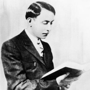 Wallace Fard Muhammad reading a book with a serious face while wearing a long sleeve under a necktie and coat