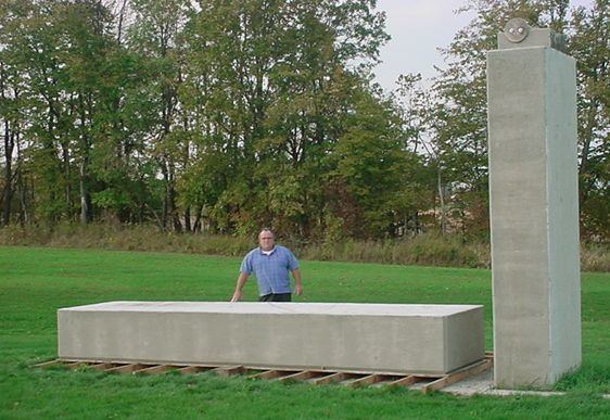 Wally Wallington posing with one of his monolith construction in an open field and wearing a sky blue shirt.