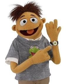 Walter with a smiling face while pointing at his watch, wearing a gray shirt with a print of a green frog over a white polo shirt and brown pants.