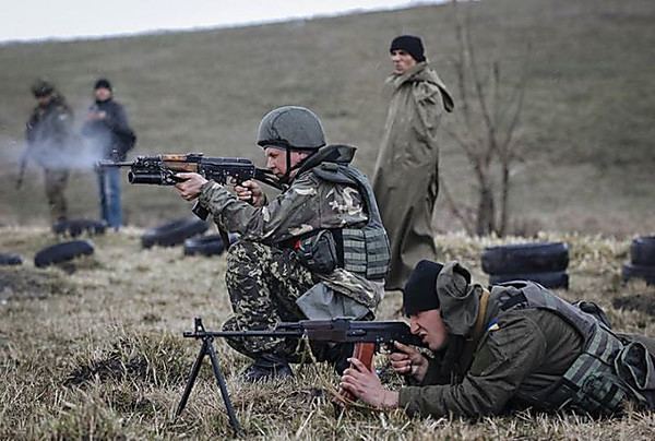 Two Ukrainian soldiers firing targets with their rifles. The soldier at the center is in a sitting position and the soldier on the right side is lying on the ground with serious face while they are in the mountain with one other soldier and two men in the background. The soldier at the center is wearing Army Combat Uniform, gray helmet, gray vest, and black shoes. The soldier on the right side is wearing a dark gray hooded jacket, gray vest, and black beanie. The soldier in the background is also wearing an Army Combat Uniform, the man next to him is wearing a black beanie, a black jacket, and denim pants while the other man is wearing a black beanie and greenish-gray cloak