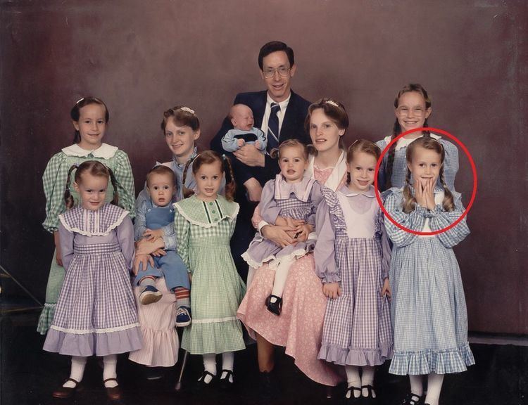 A family picture of Warren Jeffs (center, holding an infant), who has black hair and wearing eyeglass, white polo with a black necktie under a black coat. They are smiling together with his wife (4th from the right), who is carrying a girl, and she is wearing a pink retro style dress and with 10 children who wear smocked retro-style dresses and black shoes, and on the front right, with a red encircle is Rachel