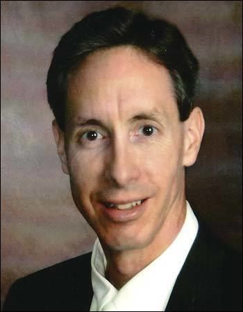 Warren Jeffs is smiling and has black hair, wearing a white polo under a black coat.