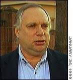 Webster Hubbell talking in a serious face with his white hair wearing a blue inner collared shirt and black coat.