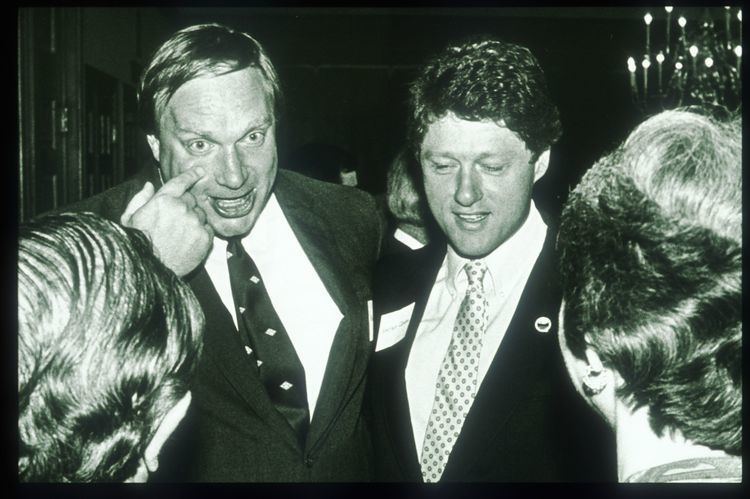 Webster Hubbell with then-Governor Bill Clinton (from center left to center right) are both smiling and talking to two women in from of them, attending a social function in Little Rock, Arkansas in 1986. Webster is touching his face with his finger and looking at someone wearing a diamond print necktie on a white collared shirt under a black coat. And Bill with a pin on his left coat is wearing a light-colored necktie with print, a white collared shirt and a black coat.