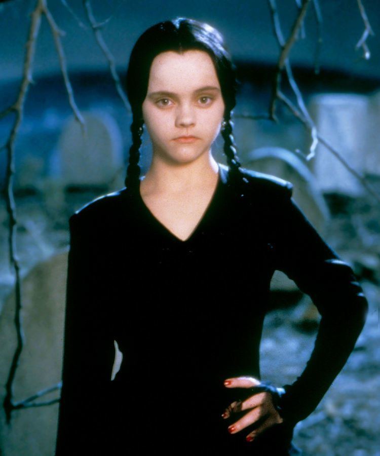 In a graveyard with tree branches and gravestones at the back, Wednesday Addams is serious, standing, with her left hand on her waist, has black hair and red fingernails wearing a black long sleeve v-neck dress,