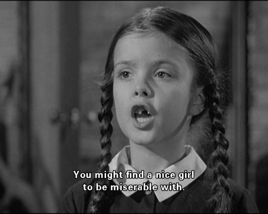 In black and white Wednesday Addams  is serious, speaking, mouth open. With her missing teeth has, black hair wearing a white collar black dress, with a subtitle written in front “You might find a nice girl to be miserable with.”
