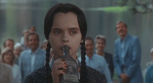 In a room with people at the back wearing formal attires, Wednesday Addams is serious, holding a black bottle with skull and poison label on with her right hand, has black hair, red fingernails wearing a white collar black dress.