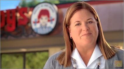 Wendy Thomas smiling outside of Wendy’s restaurant, a scene from a new Wendy’s Advertisement. She has a blonde hair, wearing round earrings, a pebble necklace, and a white polo long sleeve under a gray cardigan