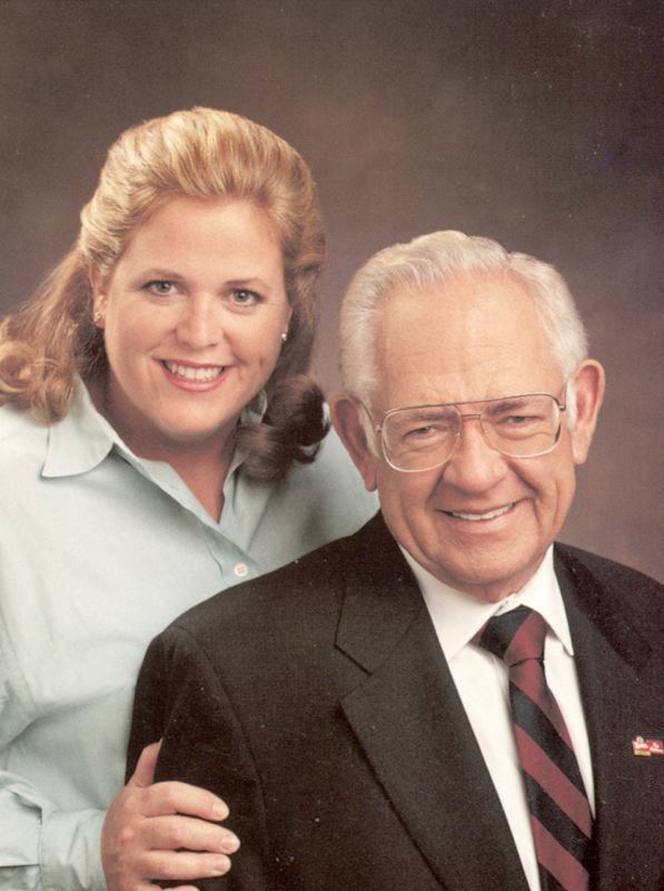 Wendy Thomas (left) smiling while touching Dave Thomas’s (right) arm in an old photograph.  Wendy had blonde curly short hair, wearing studs earrings and a white polo long sleeve. Dave had a white hair wearing eyeglasses and a white polo sleeve with a black and red necktie under a black coat