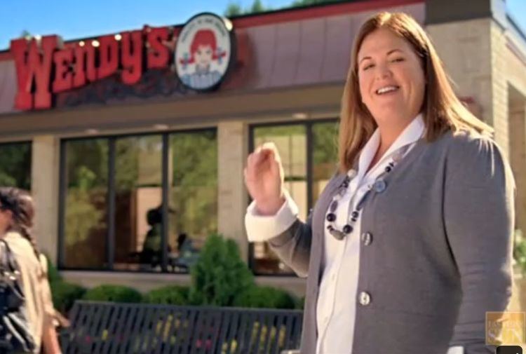 Wendy Thomas smiling outside of Wendy’s restaurant, a scene from a new Wendy’s Advertisement. She has a blonde hair, wearing a long pebble necklace, and a white polo long sleeve under a gray cardigan