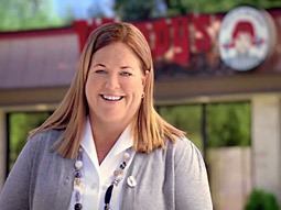 Wendy Thomas smiling outside of Wendy’s restaurant, a scene from a new Wendy’s Advertisement. She has a blonde hair, wearing round earrings, a pebble necklace, and a white polo long sleeve under a gray cardigan