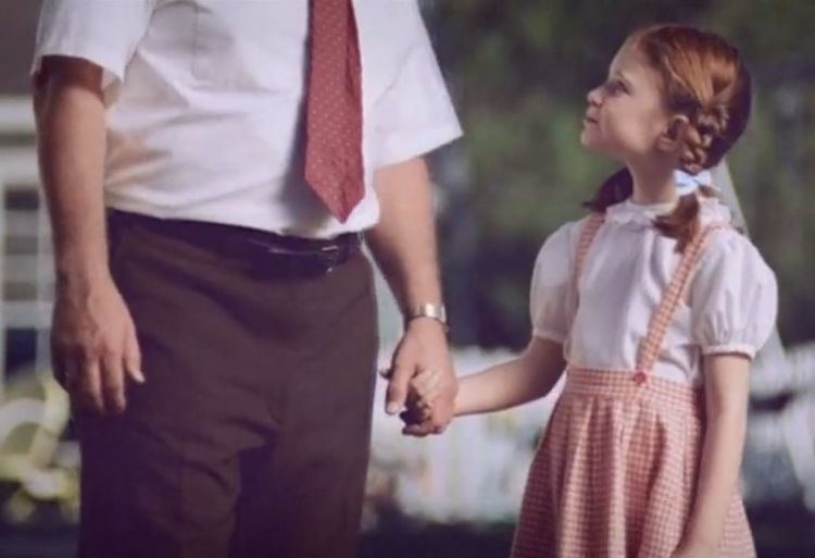 A little girl portrays as little Wendy Thomas (right) smiling while looking at and holding a man’s left hand in a scene from a new Wendy’s Advertisement. She had a braided copper blonde hair with a light blue ribbon wearing a white top under a stripe red jumper dress while the man beside her wears a watch in his left hand, white polo inserts with a red necktie, and brown pants with a black belt.