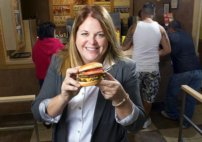 Wendy Thomas smiling while holding a burger with both hands inside of a restaurant with 3 customers at her back. She has blonde hair, wearing a round earring, a bracelet, and a white polo long sleeve under a gray coat
