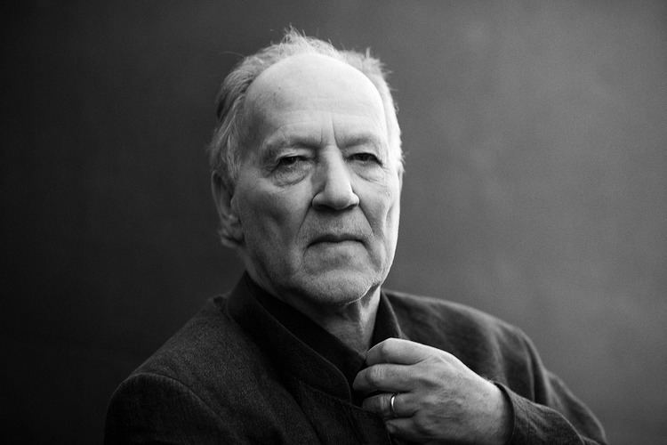 Werner Herzog posing with his hand near his neck and wearing a thick black coat.