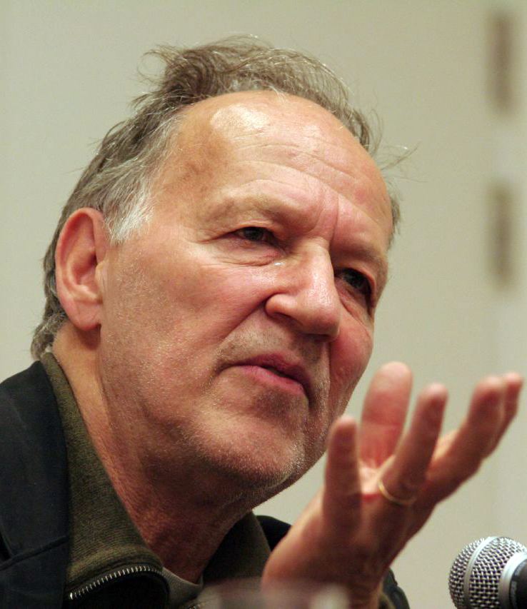 Werner Herzog talking during an interview with a bit of facial hair and wearing a dark green shirt and a black coat.