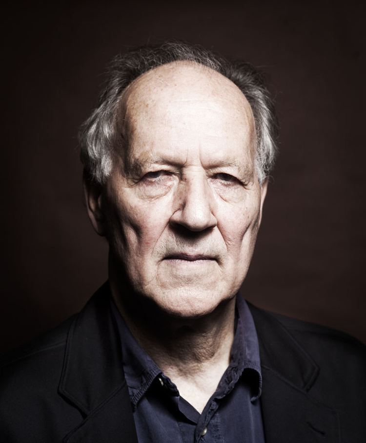 Werner Herzog posing and wearing a dark blue polo shirt underneath a striped black coat.