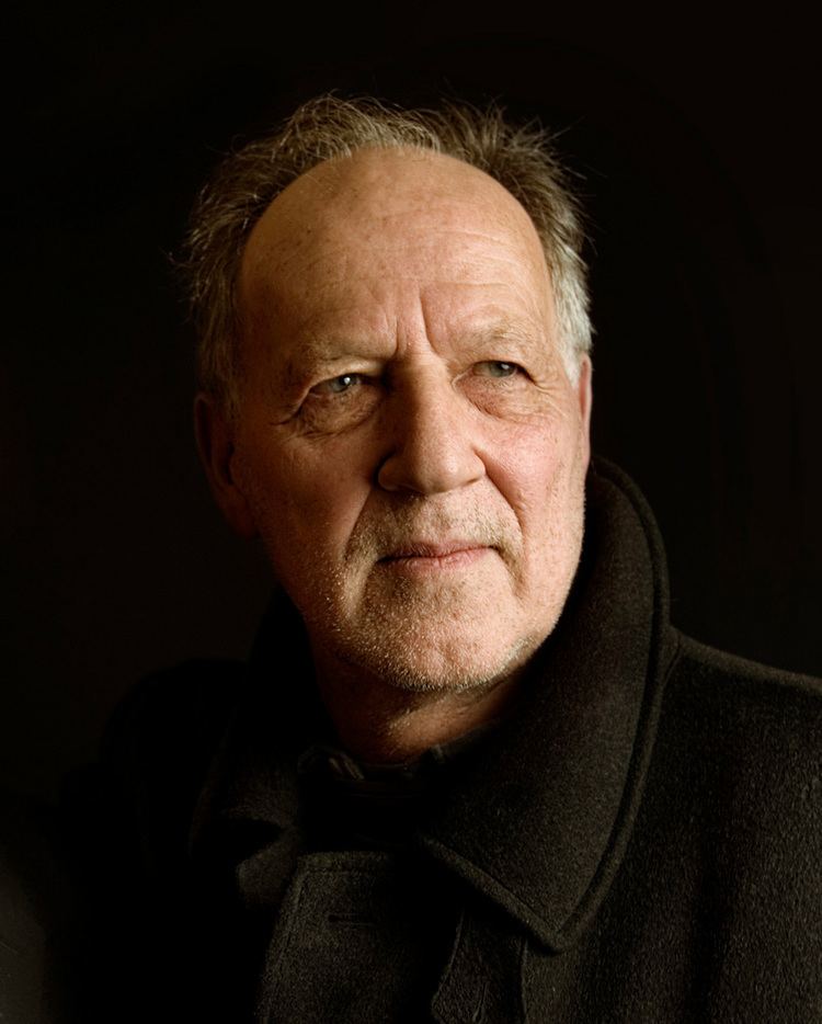 Werner Herzog posing to his side in a black background and wearing a black coat.