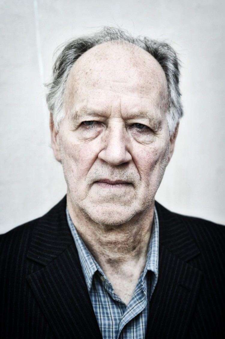 Werner Herzog posing and wearing a sky blue checkered polo shirt underneath a striped black coat.