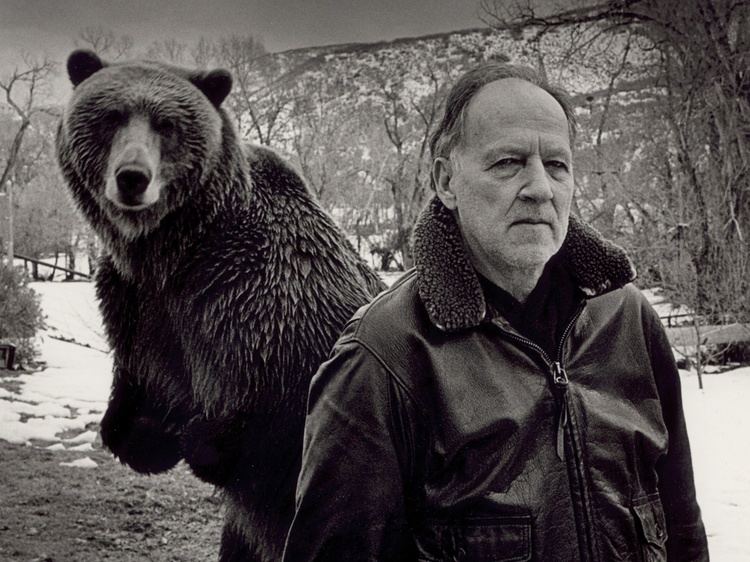 Werner Herzog posing in the snow with a bear behind him and wearing a black winter coat.