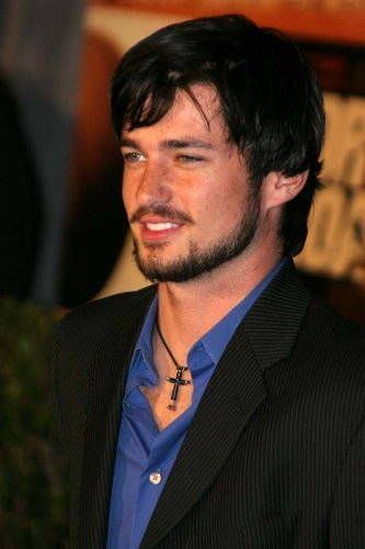 Wes Brown smiling while wearing black coat, blue long sleeves and cross necklace