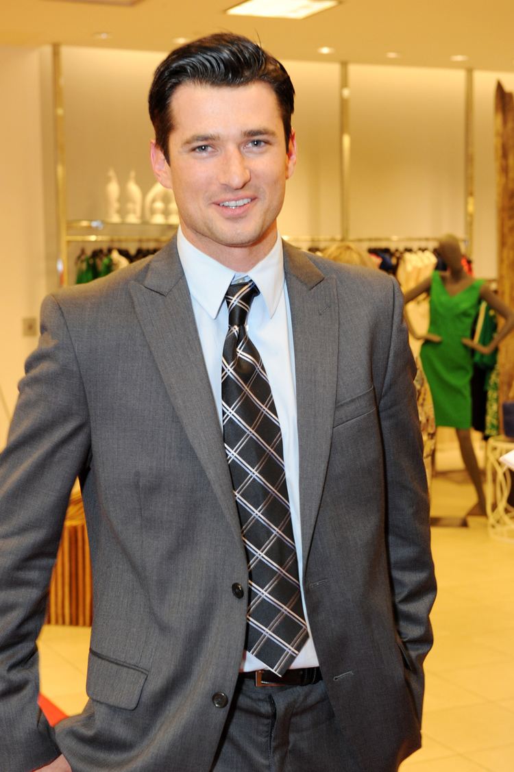 Wes Brown smiling while wearing gray coat, white long sleeves and checkered neck tie