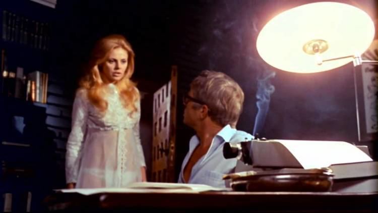 Britt Ekland and Hardy Krüger are talking to each other. Britt is wearing a white long sleeve dress and Hardy is wearing a light blue long sleeve in a scene from the 1971 horror film, What the Peeper Saw