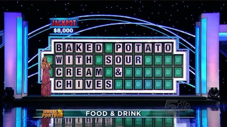 Vanna White is smiling while clapping her hands in front of the Wheel of Fortune's word board with "Baked Potato With Sour Cream & Chives" words on it. Vanna with blonde hair and wearing a multi-colored spaghetti dress.