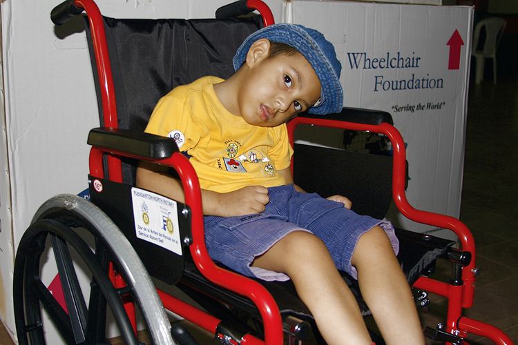 A disabled child sitting in a red and black wheelchair with a Wheelchair Foundation box in the background, he wears a blue denim hat, yellow shirt, and blue denim shorts