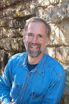 William Cronon smiling with mustache and beard while wearing eyeglasses and blue long sleeves