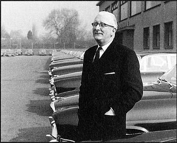 William Lyons standing beside the cars while wearing a coat, long sleeves, necktie, and eyeglasses