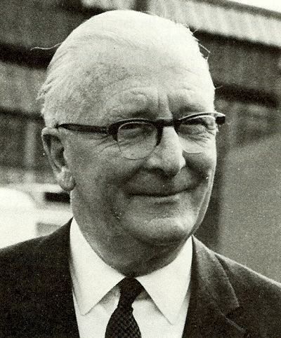 William Lyons with a tight-lipped smile while wearing a black coat, long sleeves, necktie, and eyeglasses