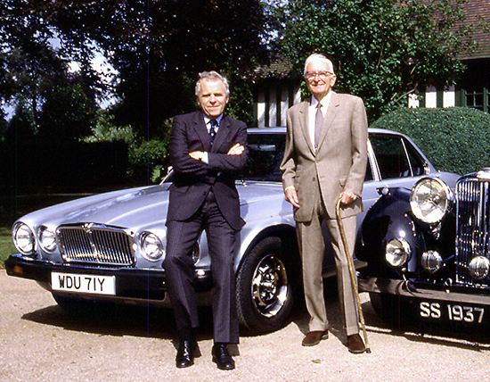 William Lyons smiling with John Egan and posing beside the Jaguar car while he is wearing a beige coat, beige pants, and white long sleeves