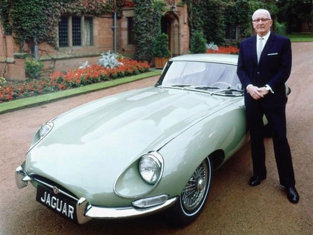 William Lyons standing beside the Jaguar E-Type while wearing a black coat, white long sleeves, necktie, black pants, and black shoes