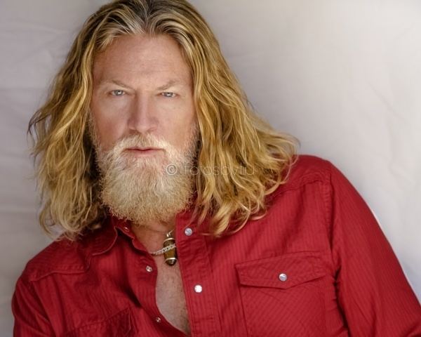 William Shockley posing with longer blonde hair and thick beard while wearing a red long-sleeved buttoned shirt.