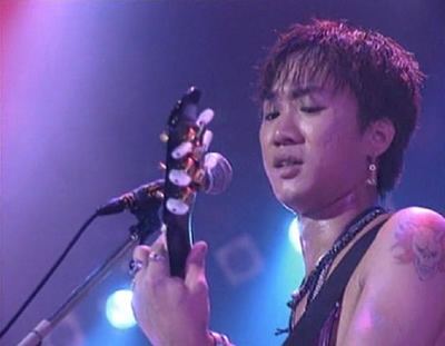 Wong Ka Kui topless and with a sad face while playing guitar with a microphone in front of him.