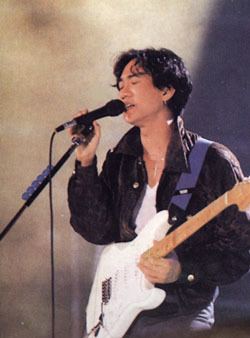 Wong Ka Kui with closed eyes while singing and playing guitar and holding a microphone, wearing a black jacket over a white shirt.