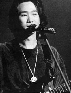 Wong Ka Kui singing and playing guitar with a microphone in front of him, wearing a necklace, and a black long sleeve shirt.