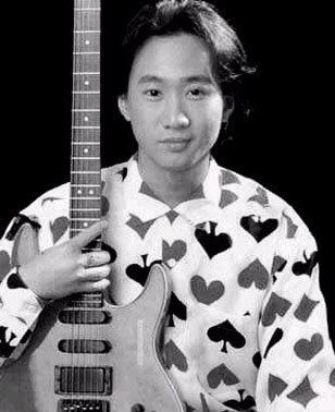 Wong Ka Kui with a tight-lipped smile while holding a guitar and wearing a white long sleeve shirt with a heart design.