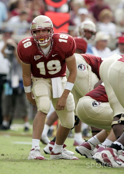 Together with his teammates, Wyatt Sexton is serious, his right arm is behind his legs, his left knee is bent, and he is wearing a brown-red helmet, a red padded jersey with a number 19 printed on the center and on both of his shoulders, a Nike logo on his left chest, and a minimal design on his right chest, both of his hands are wearing white turf tape, he is also wearing an off white shorts with a Nike logo on right, with thigh pads and knee pads with a football towel at the center, white socks paired with red and white shoes.