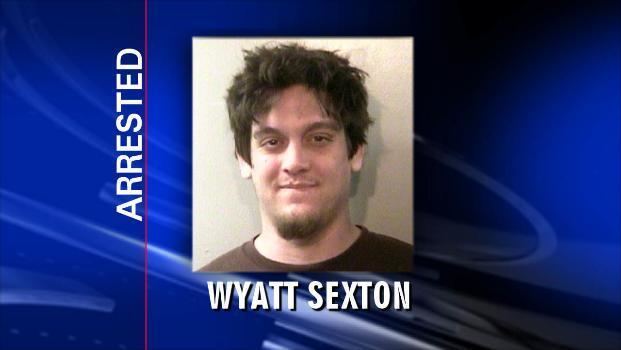On a report, is a picture of Wyatt Sexton being arrested, he is smiling, has brown hair, a mole on his right cheek, a mustache, and a beard, and he is wearing a black shirt.