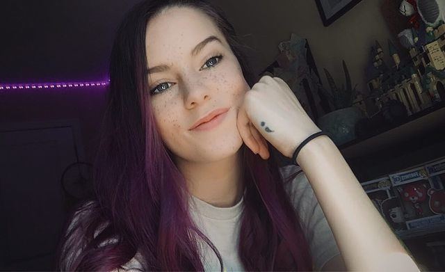 XxiiRavela smiling with her hand on her face, with long purple hair, and wearing an off-white shirt.