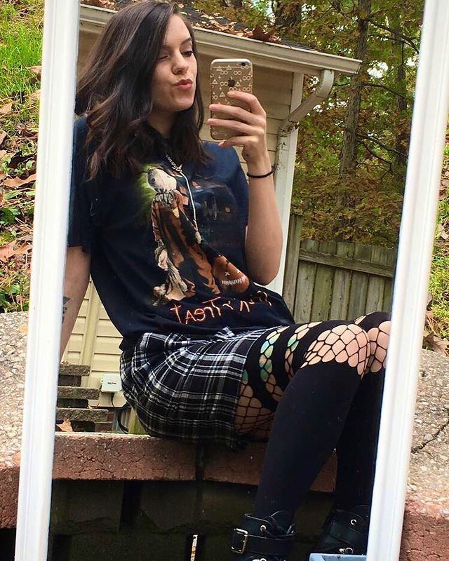 XxiiRavela pouting her lips while holding her phone, doing a selfie in front of a mirror while sitting on the stairs, with wavy short hair, wearing a blue shirt, a checkered black and white skirt, black stockings, and black shoes.