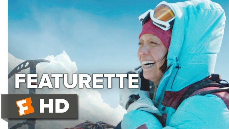 Naoko Mori as Yasuko Namba smiling happily during her mountain climbing with an ice axe and a mountain of ice in the background from a scene of a real-life film, "Everest". She is carrying a backpack with ski goggles on the top of her forehead, a red bonnet, a black and white glove, and a light blue with a red accent puffer jacket.